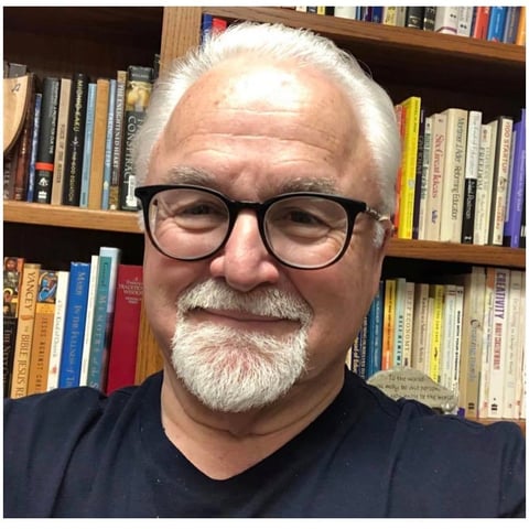 Older man with white hair and beard wearing glasses and dark shirt, smiling in front of a bookshelf